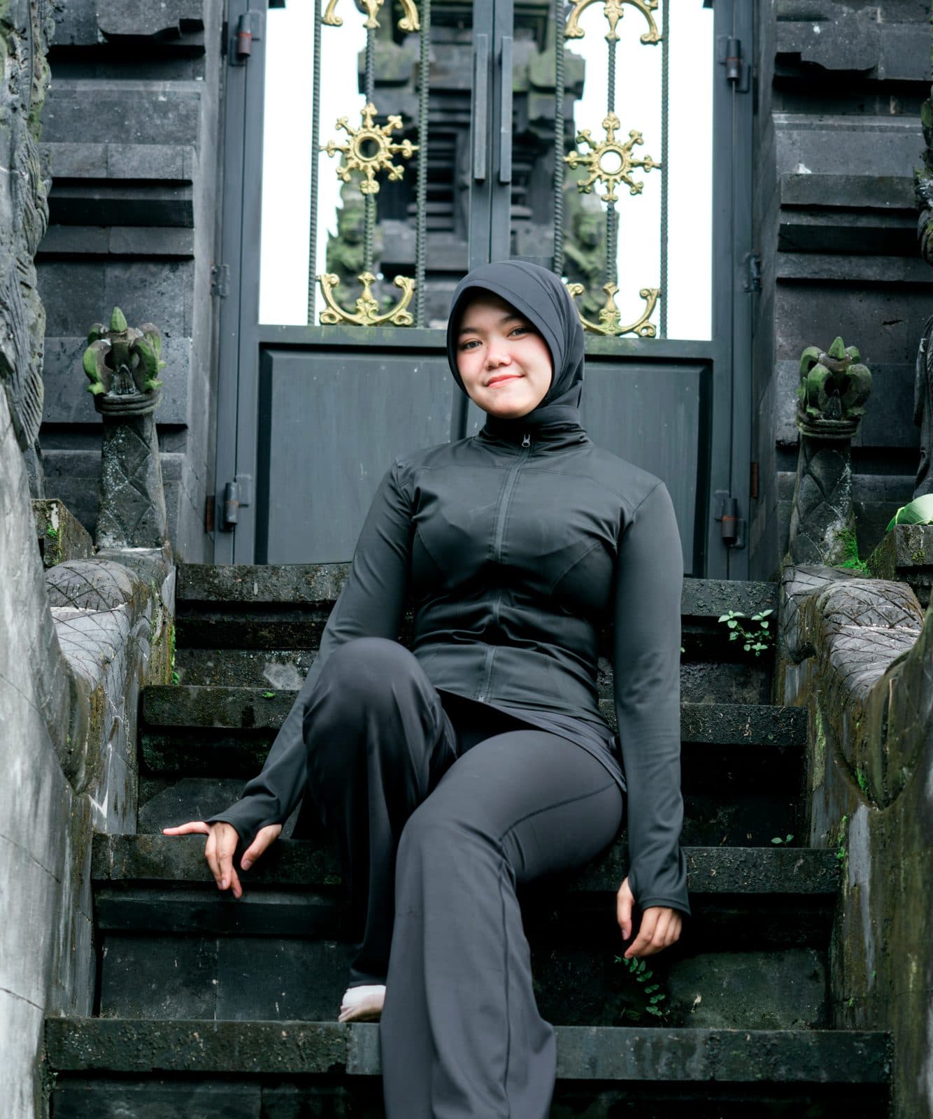 Woman wearing a black zip jacket, flared pants and sports hijab sitting on steps